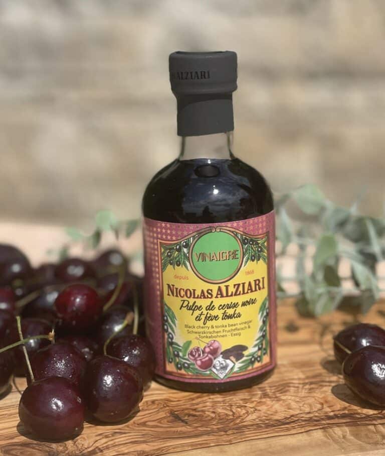 Flaska med Alziari vinäger med svart körsbär och tonkaböna (200g), placerad på ett mörkt träbord bredvid färska körsbär, hela tonkabönor och en skål med mörk choklad. Lyxig gourmetmiljö.