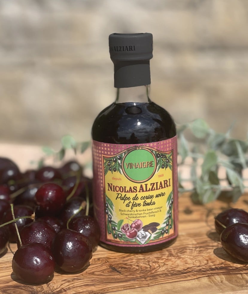 Flaska med Alziari vinäger med svart körsbär och tonkaböna (200g), placerad på ett mörkt träbord bredvid färska körsbär, hela tonkabönor och en skål med mörk choklad. Lyxig gourmetmiljö.