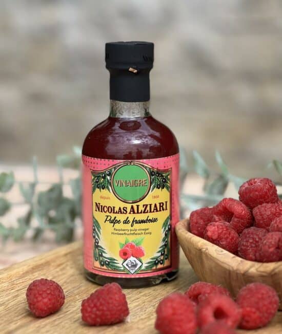 Flaska med Alziari vinäger med hallonmassa (200g), placerad på ett ljust träbord bredvid färska hallon, en salladsskål och en olivoljeflaska. Fräsch och somrig gourmetmiljö.