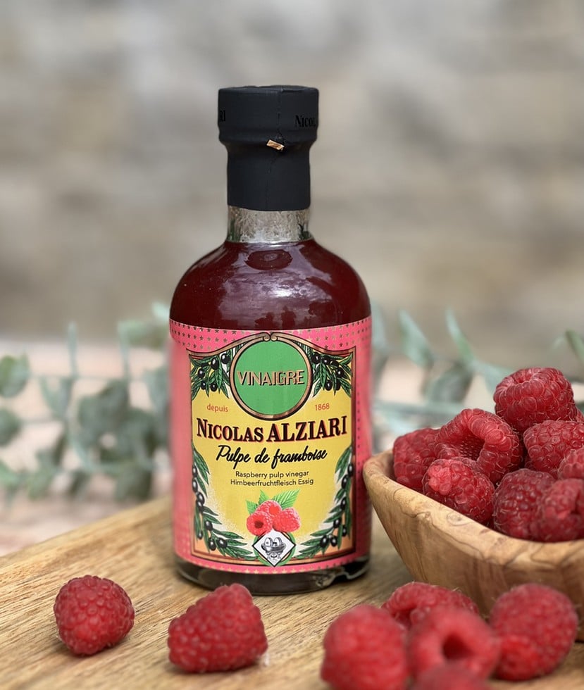 Flaska med Alziari vinäger med hallonmassa (200g), placerad på ett ljust träbord bredvid färska hallon, en salladsskål och en olivoljeflaska. Fräsch och somrig gourmetmiljö.