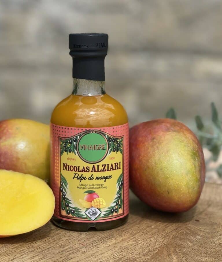 Flaska med Alziari vinäger med mangomassa (200g), placerad på ett ljust bord bredvid färska mangoskivor, lime och mynta. Ljus och exotisk matlagningsmiljö.