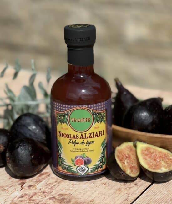 Flaska med Alziari vinäger med fikonmassa (200g), placerad på ett mörkt träbord bredvid färska, halverade fikon, en bit chèvreost och olivolja. Varm och rustik gourmetmiljö.