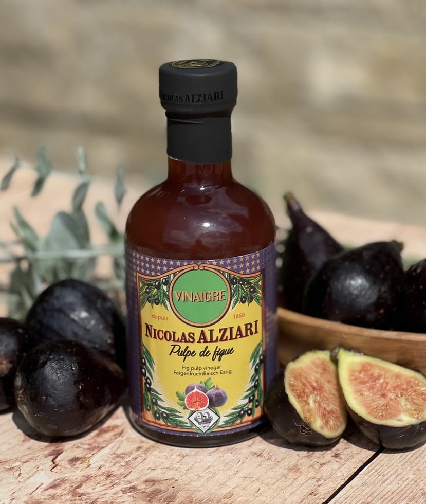 Flaska med Alziari vinäger med fikonmassa (200g), placerad på ett mörkt träbord bredvid färska, halverade fikon, en bit chèvreost och olivolja. Varm och rustik gourmetmiljö.