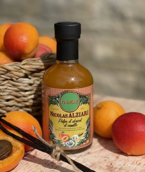 Flaska med Alziari vinäger med aprikosmassa (200g), placerad på ett ljust träbord bredvid färska, halverade aprikoser, en salladsskål och en olivoljeflaska, i soligt medelhavsljus.