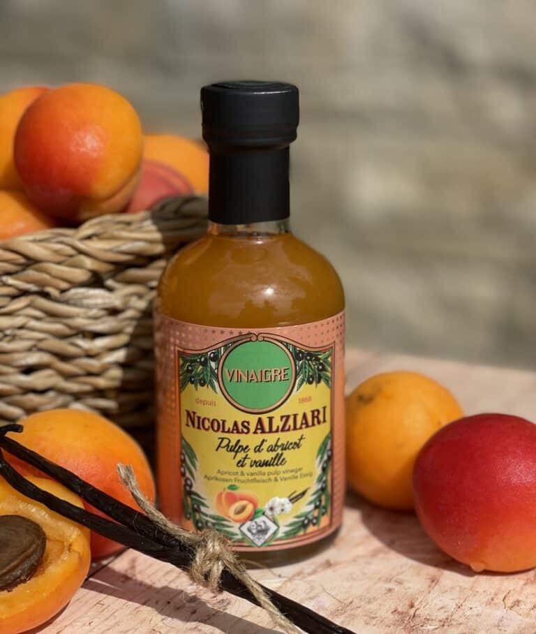 Flaska med Alziari vinäger med aprikosmassa (200g), placerad på ett ljust träbord bredvid färska, halverade aprikoser, en salladsskål och en olivoljeflaska, i soligt medelhavsljus.